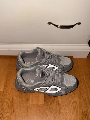 Grå Dior sneakers med CD-logga - Snygga grå sneakers från Dior med tydlig CD-logga på sidan och bak. Skorna har en chunky sula, mesh- och läderdetaljer samt grå skosnören. Perfekta för dig som vill ha en stilren och exklusiv look med streetwear-vibe.         Pris kan diskuteras vid snabb affär 