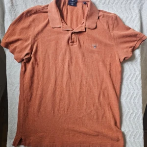 Rostbrun pikétröja från Gant - Säljer en snygg rostbrun pikétröja från Gant med klassisk krage och korta ärmar. Tröjan har diskret Gant-logga broderad på bröstet och är tillverkad i mjuk bomull. Perfekt till jeans eller shorts för en avslappnad look.