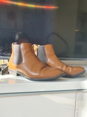 Bruna Chelsea boots i skinn - Snygga Chelsea boots i brunt skinn med elastiska svarta paneler på sidorna och låg klack. Klassisk siluett som passar till många olika stilar. Perfekta för dig som vill ha ett par stilrena boots med enkel design.