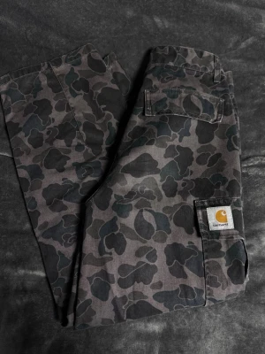 Camo cargobyxor från Carhartt - Säljer ett par camomönstrade cargobyxor från Carhartt. Byxorna har flera praktiska fickor, klassisk cargomodell och är tillverkade i slitstark bomull. Perfekta för dig som gillar streetwear och utility-vibe. Storlek M!