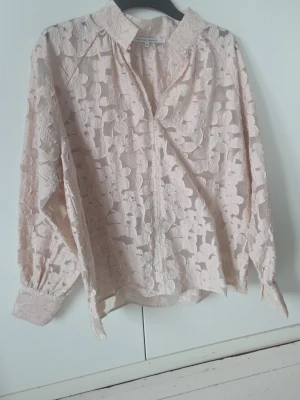 Beige spetsblus med blommönster - Söt beige (ser lite ljusrosa ut på bilderna men är beige) blus från Second Female i genombruten spets med stora blommor. Blusen har klassisk krage, knäppning framtill och långa ärmar med breda manschetter. Perfekt för dig som gillar romantisk och trendig stil.