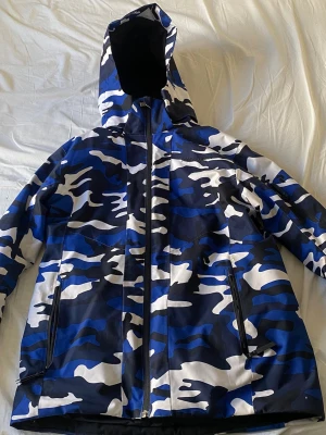 Blå camo vindjacka med huva från peakperformance - Säljer en blå, svart och vit camomönstrad vindjacka med huva. Jackan har hel dragkedja framtill, två sidofickor med dragkedja och en extra ficka på ärmen och en innerficka vid bröstet. Har använts som skidjacka. Storlek 160.