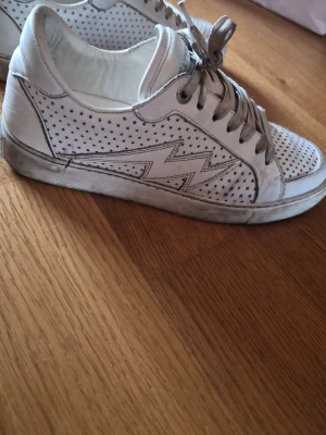 Zadig & Voltaire sneakers - Snygga vita sneakers från Zadig med små gråa stjärnor. Skorna har vita snören och perforerat stjärnmönster på tån. Tillverkade i läder med en klassisk låg profil som passar till det mesta.