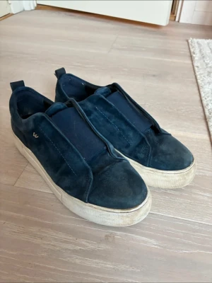 Mörkblå sneakers i mocka från K. Cobler - Snygga mörkblå sneakers från K.Cobler i mjuk mocka med bred resår över vristen istället för snörning. Skorna har vit platåsula och diskret logga på sidan. Perfekta för dig som gillar stilrena och bekväma sneakers med en twist.