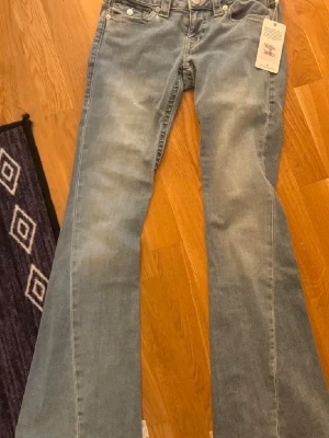 Ljusblå bootcut jeans från True Religion - Säljer ett par ikoniska True Religion jeans i modellen Joey Low Rise Flare. Jeansen är helt nya med prislappen kvar och är i den snygga ljusblå tvätten "Peak Spot".  Denna modell är känd för sin smickrande passform med låg midja och rejält utsvängda ben (flare/bell bottoms) som ger en perfekt Y2K-look.  Storlek: 25W - motsvarar XS