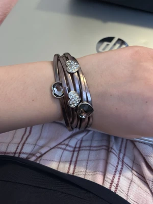 Brunt armband med hjärtan - Snyggt brunt armband som lindas flera varv runt handleden. Armbandet har silverfärgade hjärtberlocker, några med glittrande stenar och några i metall. Stängs med ett magnetlås i metall. Perfekt accessoar för en trendig look. Anledningen t att jag säljer är pga att den är inte min stil längre. Kommer inte ihåg vart jag köpte den ifrån