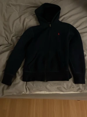 Svart hoodie från Polo Ralph Lauren - Säljer en svart hoodie från Polo Ralph Lauren med dragkedja och huva. Klassisk design med liten röd logga broderad på bröstet. Perfekt för en chill och stilren look. Tillverkad i mjukt material som känns skönt mot huden.