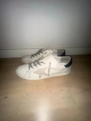 Golden Goose skor - Snygga vita sneakers från Golden Goose med grå stjärna på sidan och blå hälkappa. Skorna har grå skosnören och är tillverkade i läder med klassisk låg siluett. Perfekta för en trendig och avslappnad stil.