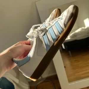 Adidas Samba vit/blå - Säljer ett par unika Adidas Samba sneakers i vitt skinn med ljusblå detaljer sparsamt använd och klassiska tre ränder på sidan.