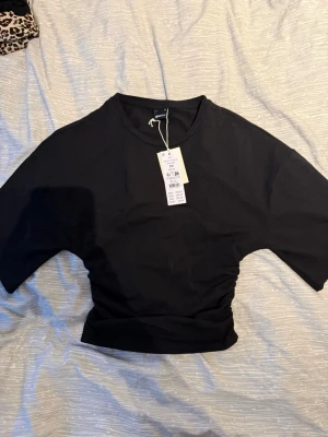 Svart croppad topp från Gina Tricot - Snygg svart croppad topp från Gina Tricot med rund halsringning och rynkade detaljer i midjan. Tillverkad i mjuk bomullsmix som känns skön mot huden. Perfekt till jeans eller kjol för en trendig look.