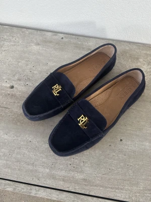 Marinblå mocka loafers Ralph Lauren - Snygga marinblå loafers i mocka från Ralph Lauren med guldfärgad RL-logga på ovansidan. Skorna har en klassisk form, bruna innersulor och gummisula med nabbad struktur för extra grepp. Perfekta för en stilren och avslappnad look.