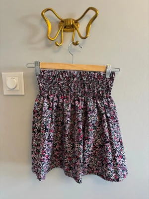 Blommig kortkjol med smock - Supersöt kortkjol med smock i midjan och blommigt mönster i rosa, lila, vitt och svart. Kjolen har en luftig passform och är perfekt för varma dagar. Tillverkad i mjukt bomullstyg som känns skönt mot huden.