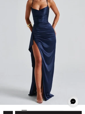 Marinblå långklänning med slits - Marinblå långklänning i satinliknande material med smala axelband och korsad rygg. Klänningen har en hög slits på ena sidan och snörning i ryggen för en snygg passform. Säljer då den är för liten i bysten. Den är aldrig använd bara provat på den.