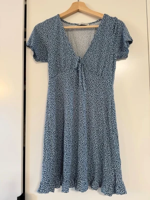 Blå blommig kortklänning med knyt - Söt blå kortklänning med små vita blommor, v-ringning och knyt framtill. Klänningen har korta ärmar och volangkant nertill för en extra feminin touch. Perfekt för varma dagar och enkel att styla med sneakers eller sandaler.