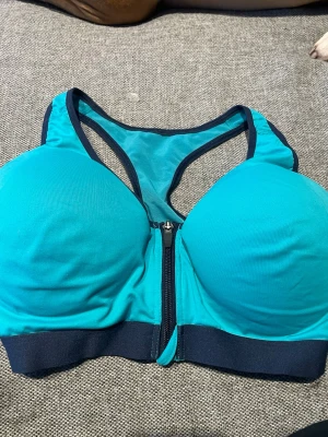 Turkos sport-bh med dragkedja fram - Sport-bh från Crivit i turkos med mörkblå detaljer och dragkedja framtill. Bh:n har vadderade kupor, bred resår under bysten och meshpartier i ryggen för extra ventilation. Perfekt för träning och ger bra stöd.
