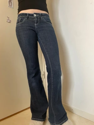 Mörkblå bootcut jeans med ljusa sömmar st 32 - Säljer ett par mörkblå bootcut jeans med vita kontrastsömmar och klassiska fem fickor. Jeansen har låg midja och utsvängda ben, perfekta för en trendig och avslappnad look. Mått finns i bilderna. 