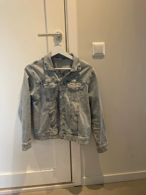 Ljusblå jeansjacka från Lindex - Snygg ljusblå jeansjacka från Lindex med klassisk krage, två bröstfickor med lock och knappar framtill. Perfekt att slänga över en hoodie eller t-shirt för en chill look. Passar dig som gillar enkel och tidlös stil.