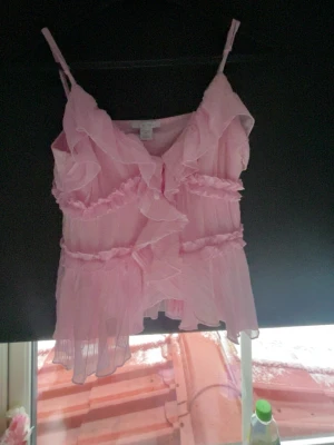 Rosa volangigt topp  med spets - Söt topp  i ljusrosa med tunna axelband och volangdetaljer. Tillverkat i ett transparent, lätt och luftigt material med spets och rysch längs hela plagget. Perfekt för dig som gillar romantisk och feminin stil på party plagg 