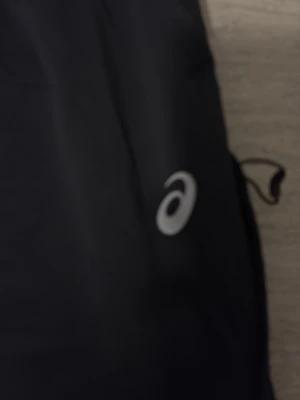 Svarta träningsbyxor från Asics - Säljer ett par svarta träningsbyxor från Asics med smal passform och dragkedjor vid fickorna. Byxorna har en diskret logga på låret och är tillverkade i ett lätt och stretchigt syntetmaterial, perfekta för löpning eller gym.