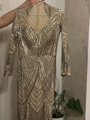 Glittrig långklänning i guld - Säljer en superglittrig långklänning i guld med långa ärmar och snyggt geometriskt mönster. Klänningen har en djup v-ringning och är täckt av paljetter som verkligen fångar ljuset. Perfekt för dig som vill sticka ut på bal eller fest!