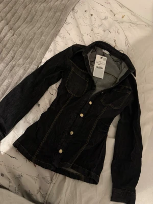 Svart figursydd jeansjacka med knappar - Säljer en svart figursydd jeansjacka från Zara med klassiska guldiga knappar och kontrasterande sömmar. Jackan har två bröstfickor med lock och lång ärm. Perfekt för dig som vill ha en stilren och snygg look. Lapparna fortfarande på aldrig använd den är från Zara. Storlek xs.