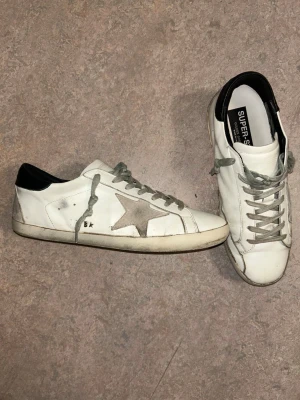 Golden Goose vita sneakers med stjärna - Golden Goose sneakers i vitt läder med grå mockadetaljer och en ikonisk stjärna på sidan. Skorna har svarta hälkappor, grå skosnören och slitna detaljer för en cool, avslappnad look. Perfekta för dig som gillar streetstyle och unika detaljer.   Jag kan inte 100 procent säga om de är äkta eftersom att de är andrahand. Men den tidigare ägaren hävdade att de var äkta samt medföljde äkthetsbevis! Tveka inte att höra av dig vid intresse☺️