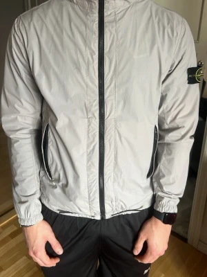 Ljusgrå vindjacka från Stone Island - Snygg ljusgrå vindjacka från Stone Island med svart dragkedja framtill och två fickor med dragkedja. Jackan har den klassiska Stone Island-loggan på vänster ärm och elastiska muddar vid handlederna. Tillverkad i lätt och tunt material, perfekt för lager-på-lager.