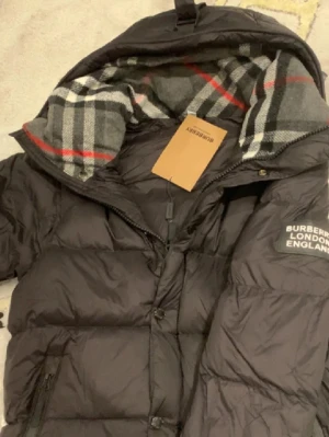 Svart pufferjacka från Burberry - Säljer en svart pufferjacka från Burberry med klassiskt rutigt foder i grått, vitt och rött. Jackan har huva, dragkedja och tryckknappar samt en patch med Burberry London England på ärmen. Perfekt för kalla vinterdagar.