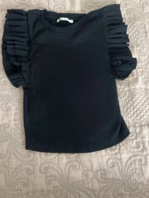 Svart topp med volangärmar - Trendig svart topp från ZARA med korta volangärmar som ger en cool och unik look. Toppen har rund halsringning och är gjord i ett mjukt, ribbat material. Perfekt för dig som vill sticka ut lite extra.