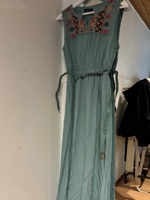 Mint långklänning med broderi - Långklänning i ljusgrön med broderade detaljer i flera färger vid halsringningen. Klänningen är ärmlös och har ett smalt bälte i midjan. Bältet har några defekter, inget som syns!💕