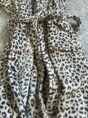 Leopardmönstrad mjuk morgonrock - Supermjuk morgonrock från Åhléns i leopardmönster. Beige botten med svarta och grå fläckar, knytskärp i midjan och praktisk ficka framtill. Perfekt för att mysa runt hemma i. Tillverkad i fluffigt, värmande material.