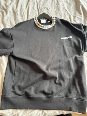 Svart sweatshirt med rutigt mönster - Snygg svart sweatshirt med klassiskt beige, svart och rött rutigt mönster i halsringningen. Tröjan har en rak passform och ribbade muddar vid ärmslut och nederkant. Perfekt för dig som gillar stilrena plagg med en twist.