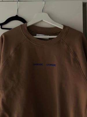 Brun sweatshirt från Samsøe Samsøe - Snygg brun sweatshirt från Samsøe Samsøe med rund halsringning och diskret blå logga broderad framtill. Tröjan har en avslappnad passform och ribbade muddar vid hals, ärmslut och nederkant. 