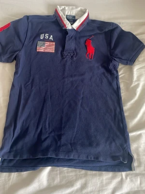 Blå Polo Ralph Lauren rugbytröja - Säljer en mörkblå rugbytröja från Polo Ralph Lauren med vit krage och röda detaljer. Tröjan har USA-flagga och texten 'USA' broderat på bröstet samt den klassiska röda polospelaren. Kortärmad och i bomull, perfekt för en sportig look.