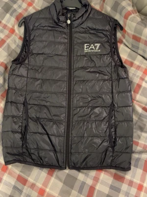 Svart dunväst från EA7 Emporio Armani - Snygg svart dunväst från EA7 Emporio Armani med quiltad design och hög krage. Västen har dragkedja framtill, två sidofickor och en vit logga på bröstet. Perfekt för lager på lager och streetwear-stil.