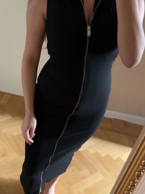 Svart klänning med dragkedja - Snygg svart klänning i figurnära modell med hel dragkedja framtill. Klänningen är ärmlös och har en stilren, modern look som passar perfekt för dig som gillar minimalistisk stil.