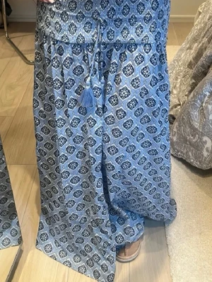 Blå mönstrad långkjol med snörning - Säljer en blå långkjol med snyggt grafiskt mönster och dekorativ snörning med tofsar i midjan. Kjolen har en avslappnad passform och är perfekt för varma dagar. Materialet känns lätt och luftigt. Strlk 158/164