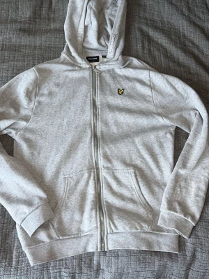 Grå hoodie från Lyle & Scott - Snygg ljusgrå hoodie från Lyle & Scott med dragkedja, huva och två fickor framtill. Klassisk gul logga på bröstet och ribbade muddar vid ärmar och nederkant. Tillverkad i mjuk bomullsblandning som känns skön mot huden.