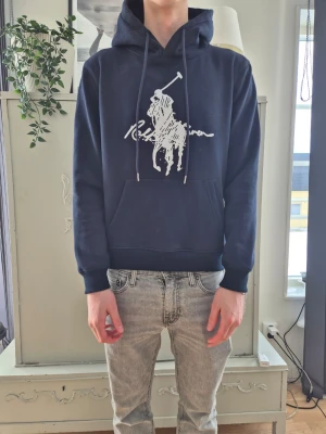 Mörkblå hoodie från Ralph Lauren - Snygg mörkblå hoodie från Ralph Lauren med stor vit logga och polospelare på bröstet. Klassisk känguruficka framtill, justerbar huva med dragsko och ribbade muddar vid ärmar och nederkant. Perfekt för en avslappnad och trendig look. Storlek M, men upplever den mer som en S.