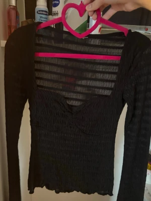 Svart mesh topp med lång ärm - Säljer en svart mesh topp med lång ärm och v-ringad urringning. Toppen är genomskinlig och har en ribbad struktur som ger en snygg och trendig look. Perfekt att styla lager på lager eller med ett linne under. Figurnära passform och vågig kant nertill. Aldrig använd.