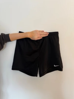 Svarta träningsshorts från Nike XS - Säljer ett par svarta träningsshorts från Nike med elastisk midja och klassisk vit logga på benet. Shortsen är tillverkade i ett lätt och svalt polyestermaterial, perfekta för sport eller gym. Är i barnstorlek XL men skulle säga att det motsvarar en storlek XS.