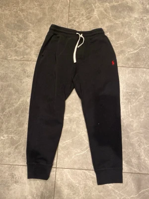 Svarta mjukisbyxor från Polo Ralph Lauren - Svarta mjukisbyxor från Polo Ralph Lauren med vit dragsko i midjan och röd logga broderad på vänster sida. Byxorna har muddar vid bensluten, sidofickor och en bakficka. Tillverkade i mjukt material som känns skönt mot huden.