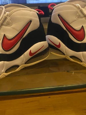 Nike Air Max Uptempo vita/röda - Nike Air Max Uptempo sneakers i vitt, svart och rött med stora röda Swoosh-loggor på sidorna och mindre Swoosh på sulan. Skorna har en chunky sula med synlig Air-enhet och röda detaljer på dragflikarna. Klassisk retrostil med högt skaft.