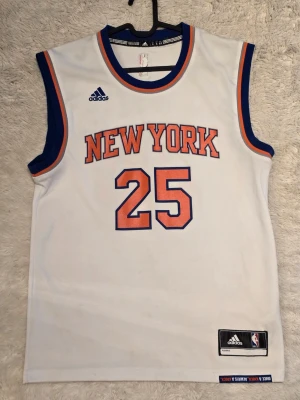 New York Knicks basketlinne Adidas - S/M.Säljer ett vitt basketlinne från Adidas med New York Knicks tryck och nummer 25 i orange och blått. Linnet har blå och orange detaljer runt hals och ärmar samt officiell NBA-logga. Perfekt för basketfans som vill ha en sportig look.