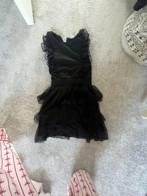 Svart volangklänning med prickigt tyll - Supersöt svart kortklänning med volanger och prickigt tylltyg. Klänningen har markerad midja, volangdetaljer på axlar och kjol samt är ärmlös. Perfekt för dig som gillar en lekfull och trendig look. Prislapp kvar.