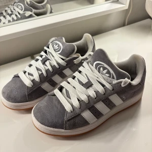 Adidas Campus grå sneakers - Säljer ett par klassiska Adidas Gazelle sneakers i grå mocka med vita ränder och vit sula. Skorna har rund tå, platt sula och breda vita skosnören. Perfekta för en stilren vardagslook.