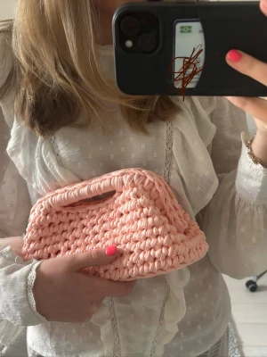 Handvirkad rosa kuvertväska - Supersöt handvirkad kuvertväska i ljusrosa med chunky struktur. Väskan har inbyggt handtag och är gjord av tjockt garn som ger en unik och trendig look. Perfekt accessoar för att lyfta vilken outfit som helst.