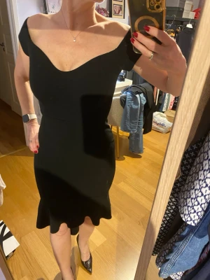 Svart offshoulder klänning - Snygg svart klänning med offshoulder och v-ringad urringning. Modellen är figurnära med volangkant nertill och kortare längd fran än bak. Köpt på Bubbleroom, aldrig använd. Storlek 38 men känns som 36.