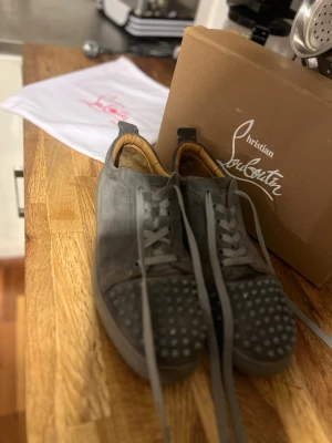 Grå Christian Louboutin sneakers - Snygga grå sneakers från Christian Louboutin med klassiska röda sulor och coola nitar på tån. Skorna är låga och har grå mocka med matchande skosnören. Perfekta för dig som vill sticka ut med exklusiv streetstyle.