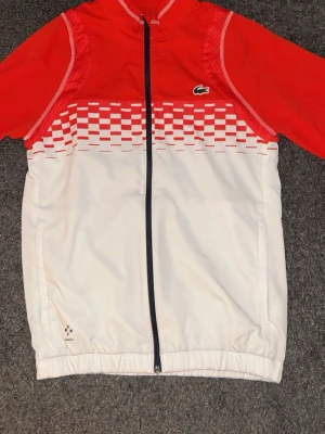 Rödvit vindjacka från Lacoste  - Säljer min unika vindjacka från Lacoste, perfekt för dig som gillar att sticka ut och ha på sig något elegant. Jackan har inga skador, fläckar eller noppor. Tveka inte att höra av dig om du är intresserad!!